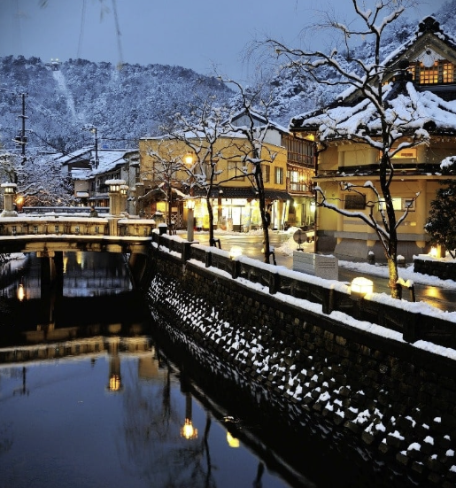 "Snow-covered street and river near Kyoto Station during winter evening with traditional buildings and warm lights reflecting on the water."