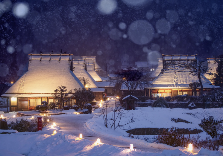 “Traditional thatched-roof houses in Kyoto illuminated at night during winter snowfall, along a serene lantern-lit walking path.”