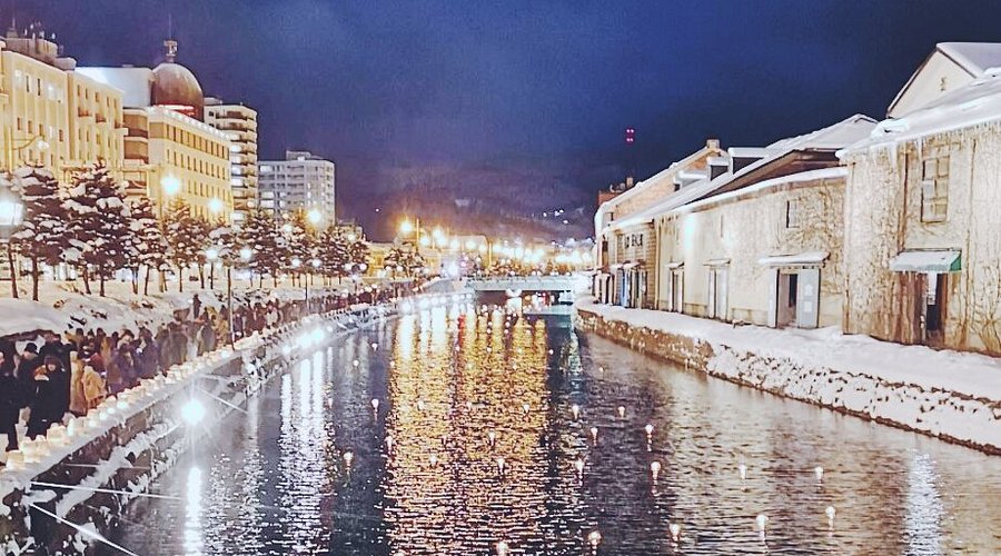 "A snowy winter night view of Otaru Canal with warm glowing lights reflecting on the water and historic buildings covered in snow."